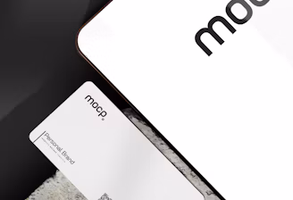A business card with 'mocp' branding placed on a concrete surface, accompanied by a modern electronic device displaying the same text. The design is minimalist, featuring white and gray tones. A QR code is present on the card alongside the text 'Personal Brand'.