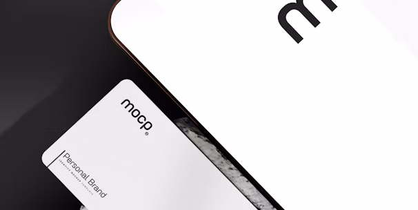 A business card with 'mocp' branding placed on a concrete surface, accompanied by a modern electronic device displaying the same text. The design is minimalist, featuring white and gray tones. A QR code is present on the card alongside the text 'Personal Brand'.
