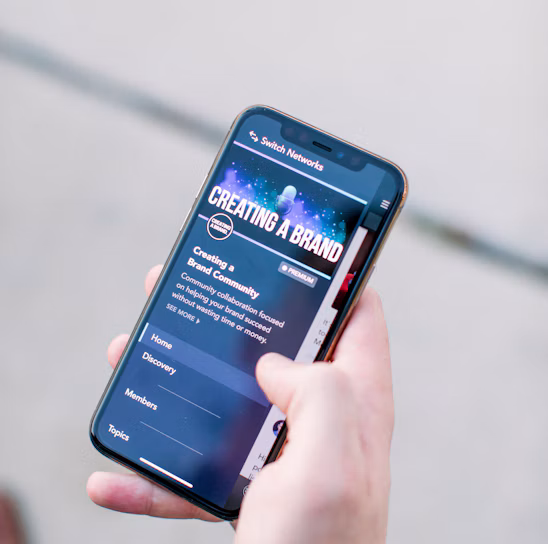 A hand holding a smartphone displaying a webpage titled 'Creating a Brand' with navigation options like Home, Discovery, Members, and Topics. The background is out of focus, providing a neutral setting.